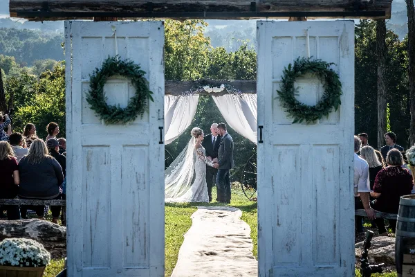 A bride and groom exchanging vows at an outdoor wedding ceremony, framed by white rustic doors with green wreaths.