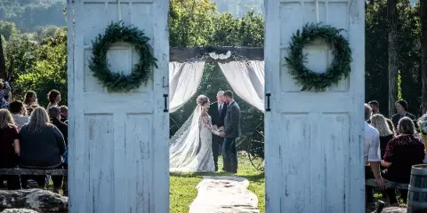 A bride and groom exchanging vows at an outdoor wedding ceremony, framed by white rustic doors with green wreaths.