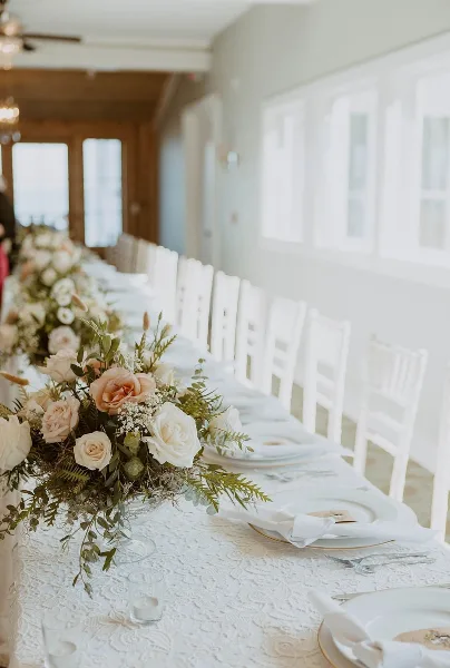 A long wedding reception table set with white chairs and a detailed floral centerpiece featuring blush and white roses with greenery.