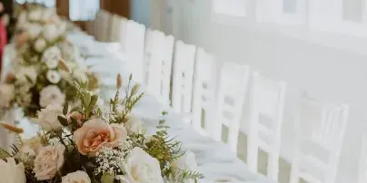 A long wedding reception table set with white chairs and a detailed floral centerpiece featuring blush and white roses with greenery.