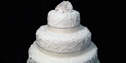 A three-tiered white wedding cake decorated with floral piping and a rose topper.