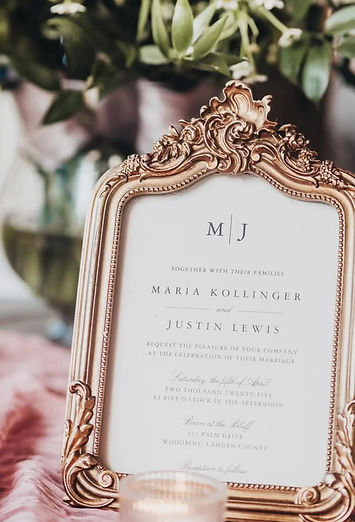 A close-up of a wedding invitation in an ornate gold frame, listing Barn at the Bluff in Woodbine, Georgia as the venue.