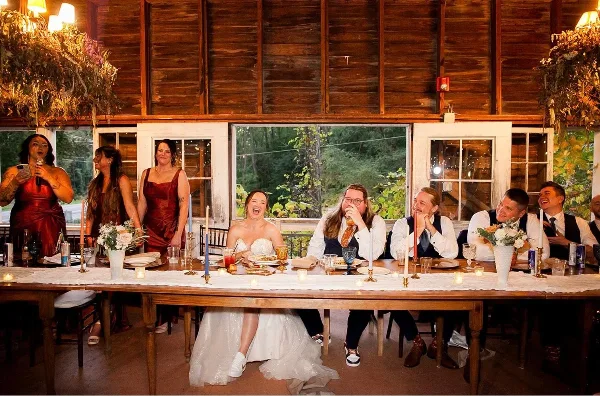 Wedding party seated at a long candlelit table with plated food in a rustic barn reception hall, bride in white dress and a woman speaking into a microphone.