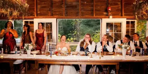 Wedding party seated at a long candlelit table with plated food in a rustic barn reception hall, bride in white dress and a woman speaking into a microphone.