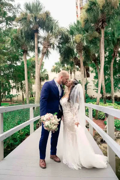A bride and groom kissing on a wooden bridge surrounded by palm trees.