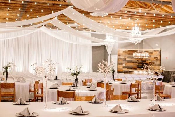 Interior of a wedding venue featuring round tables with white tablecloths, wooden chairs, and white fabric drapery hanging from a wooden ceiling.