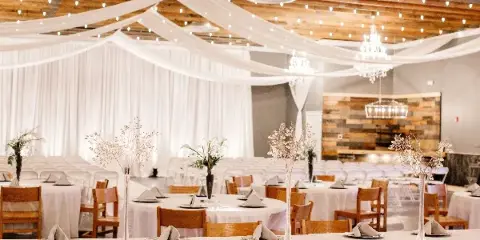 Interior of a wedding venue featuring round tables with white tablecloths, wooden chairs, and white fabric drapery hanging from a wooden ceiling.