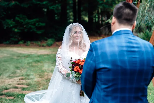 A bride in a white lace gown and veil holding a bouquet of red and orange roses, facing a groom in a blue plaid jacket during an outdoor wedding ceremony.