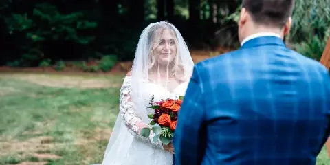A bride in a white lace gown and veil holding a bouquet of red and orange roses, facing a groom in a blue plaid jacket during an outdoor wedding ceremony.