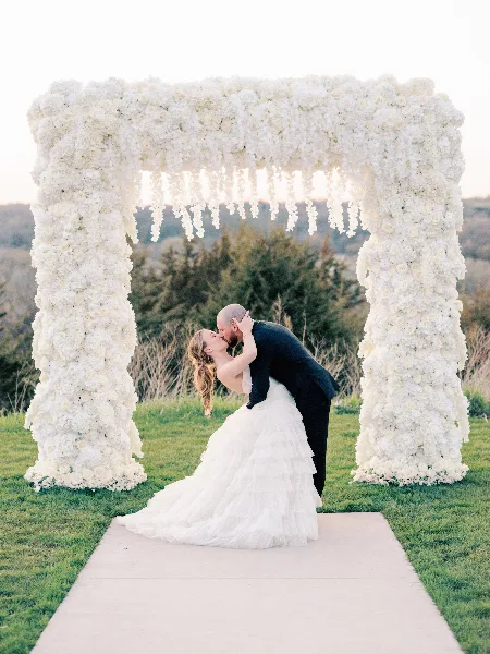 A couple kissing under a large, white floral archway at an outdoor wedding.