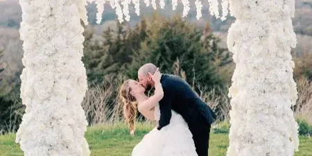 A couple kissing under a large, white floral archway at an outdoor wedding.