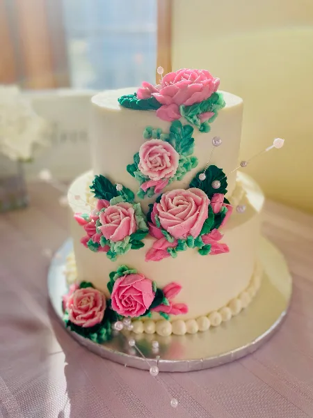 A two-tiered white wedding cake decorated with pink buttercream roses, green leaves, and pearl accents on a silver cake stand.