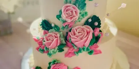 A two-tiered white wedding cake decorated with pink buttercream roses, green leaves, and pearl accents on a silver cake stand.