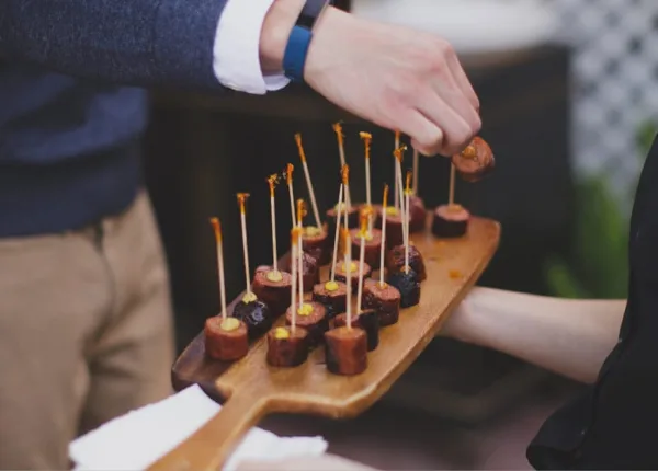 A person serving sausage roll appetizers on a wooden board at an event.