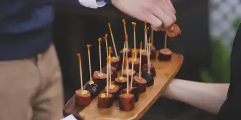 A person serving sausage roll appetizers on a wooden board at an event.
