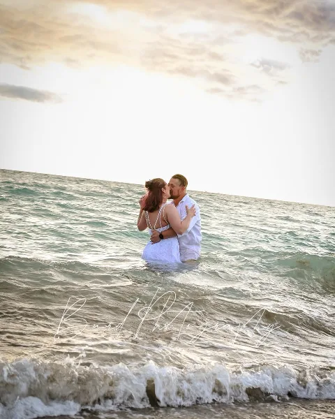 A bride and groom in white wedding attire embrace and kiss in the shallow ocean surf at a beach during golden hour.