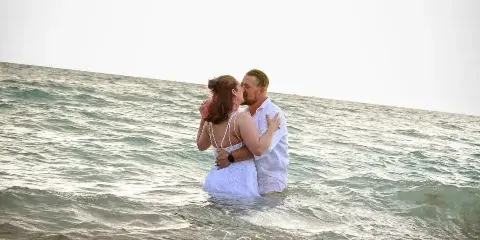 A bride and groom in white wedding attire embrace and kiss in the shallow ocean surf at a beach during golden hour.