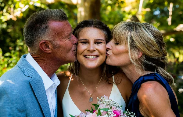 A bride in a white dress holding a bouquet of pink and white flowers is being kissed on the cheeks by her father and mother in an outdoor setting.