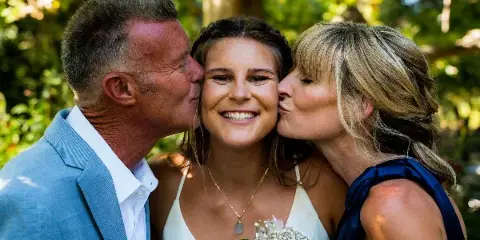 A bride in a white dress holding a bouquet of pink and white flowers is being kissed on the cheeks by her father and mother in an outdoor setting.