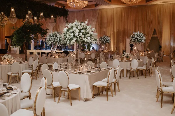 A wedding reception hall featuring round tables with white linens, gold-trimmed chairs, large white floral centerpieces, and draped fabric walls.