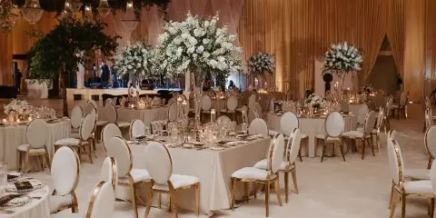 A wedding reception hall featuring round tables with white linens, gold-trimmed chairs, large white floral centerpieces, and draped fabric walls.