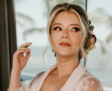 A bride with an elegant updo and professional makeup, wearing a white robe and floral hair accessory.