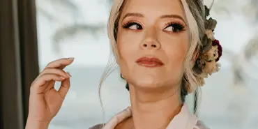 A bride with an elegant updo and professional makeup, wearing a white robe and floral hair accessory.