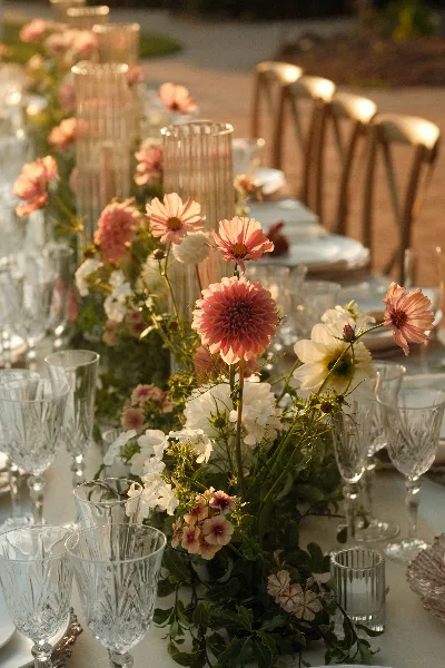 A floral table arrangement with pink dahlias and white flowers set on a dining table with glassware.