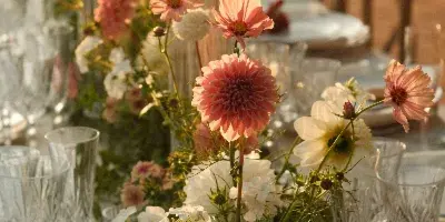 A floral table arrangement with pink dahlias and white flowers set on a dining table with glassware.