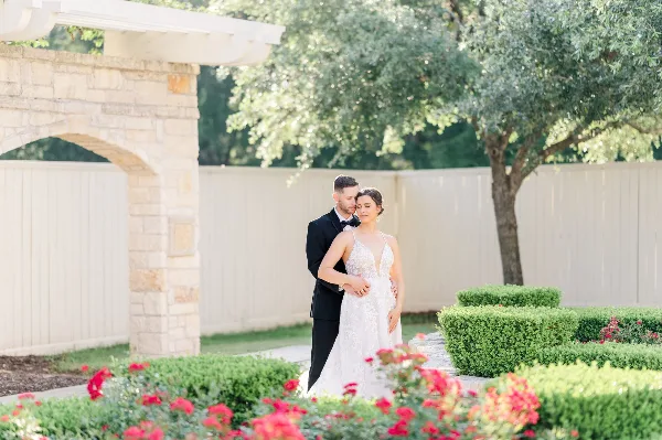 A bride and groom standing together in a garden with a stone archway and red flowers.