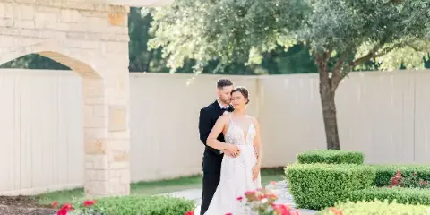 A bride and groom standing together in a garden with a stone archway and red flowers.