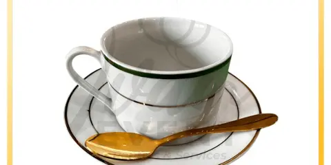 A white coffee cup with a green rim, saucer, and gold spoon set on a white background.