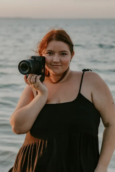 Portrait of a female photographer with red hair holding a camera near a body of water.