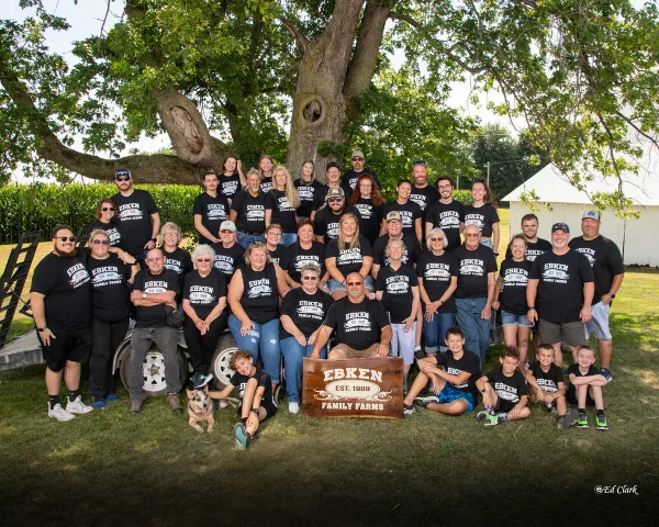 A large multi-generational family posing for a group portrait outdoors under a large tree, wearing matching black t-shirts.