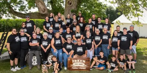 A large multi-generational family posing for a group portrait outdoors under a large tree, wearing matching black t-shirts.