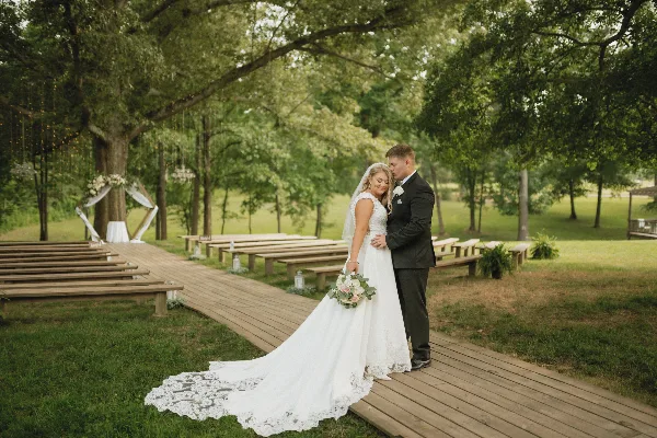 A bride and groom standing on a wooden aisle at an outdoor wedding ceremony site beneath a large oak tree.