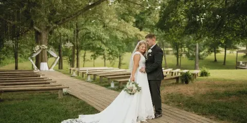 A bride and groom standing on a wooden aisle at an outdoor wedding ceremony site beneath a large oak tree.