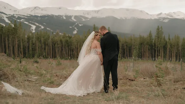 A bride and groom standing together in a field with a backdrop of pine trees and snow-capped mountains.