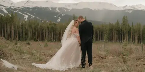 A bride and groom standing together in a field with a backdrop of pine trees and snow-capped mountains.