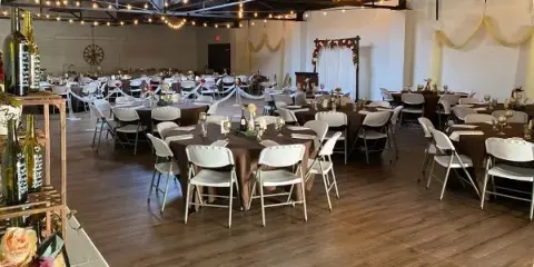 Interior view of a community hall event space with round tables, white chairs, and a floral archway.