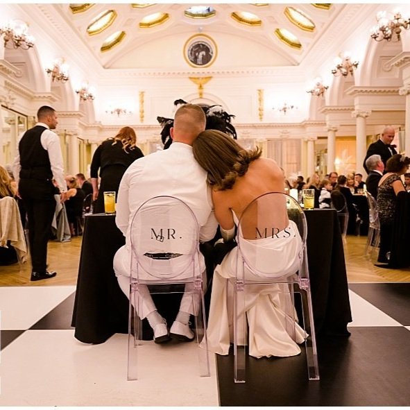 A bride and groom seated on clear chairs labeled 'Mr.' and 'Mrs.' at a wedding reception in a large, ornate ballroom.