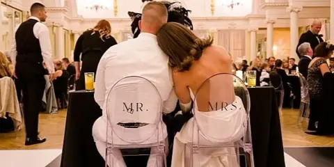 A bride and groom seated on clear chairs labeled 'Mr.' and 'Mrs.' at a wedding reception in a large, ornate ballroom.