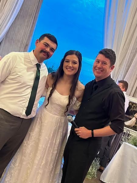 A DJ in a black shirt holding a microphone poses with a bride and groom at a wedding.