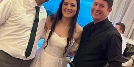 A DJ in a black shirt holding a microphone poses with a bride and groom at a wedding.
