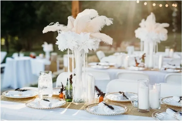 Wedding reception table setting with white feather and floral centerpieces, gold-rimmed plates, and candles under string lights.