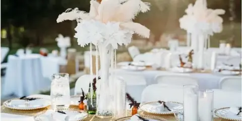 Wedding reception table setting with white feather and floral centerpieces, gold-rimmed plates, and candles under string lights.