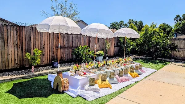 Outdoor luxury picnic setup with white umbrellas, decorated table, and floral arrangements in a backyard.