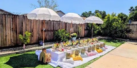 Outdoor luxury picnic setup with white umbrellas, decorated table, and floral arrangements in a backyard.