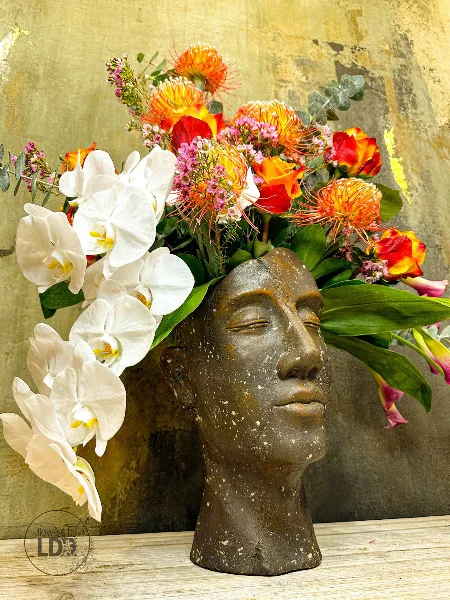 A vibrant floral arrangement with white orchids and orange proteas arranged in a sculptural head-shaped vase.