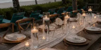 A long wooden table set for a wedding with white linens, charger plates, and LED candles in glass cylinders, situated outdoors near a beach.